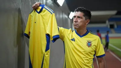 Floodlight rim light captures a determined, 30-year-old Ecuador national team coach, standing waist-up on the Estadio...