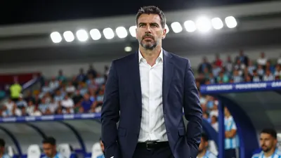A 35-year-old Argentine coach in a navy blue suit and white shirt, standing in a stadium tunnel with floodlight rim...