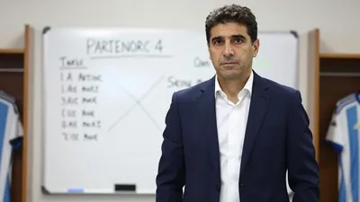 A 35-year-old Argentine football manager with short dark hair, wearing a clean navy blue suit with a white shirt...