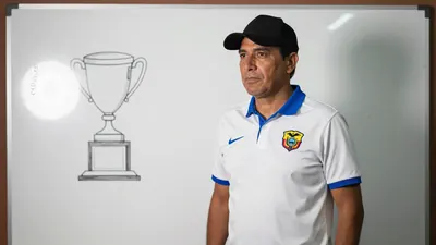 A tight crop of a 35-year-old Ecuadorian coach, wearing a white shirt with a blue collar and sleeves, and a black hat...