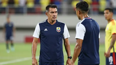 A 30-year-old Ecuador national team coach, wearing a navy blue jersey with white sleeves, stands on the sidelines of a...