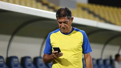 A 50-year-old Ecuador national team coach with a rugged build, wearing a plain yellow and blue polyester training top...