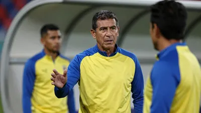 A 50-year-old Ecuadorian coach in a yellow and blue plain polyester training top, with a stern expression, stands in a...