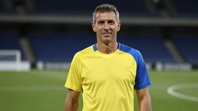 Middle-aged male club official, athletic build, waist-up, wearing a plain, unmarked training top split yellow and blue...