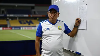 A 30-year-old Ecuadorian football coach, wearing a white jersey with blue sleeves and a blue hat, stands in front of a...