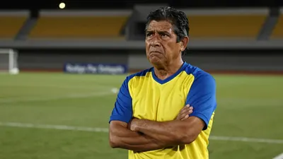 A 35-year-old Ecuadorian coach with a rugged build, stands with his arms crossed, his face set in a mix of frustration...